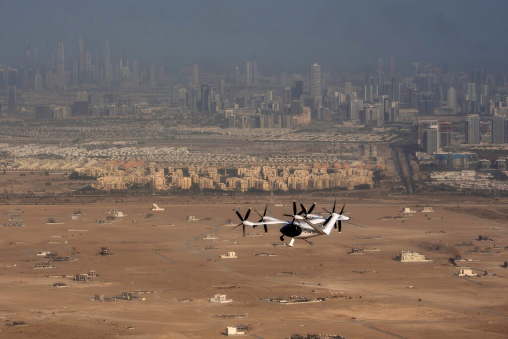 A Futuristic Looking White Aircraft Is Seen Flying Over A Brown Desert Landscape With The Skyscrapers Of A Large Modern City In Scaled