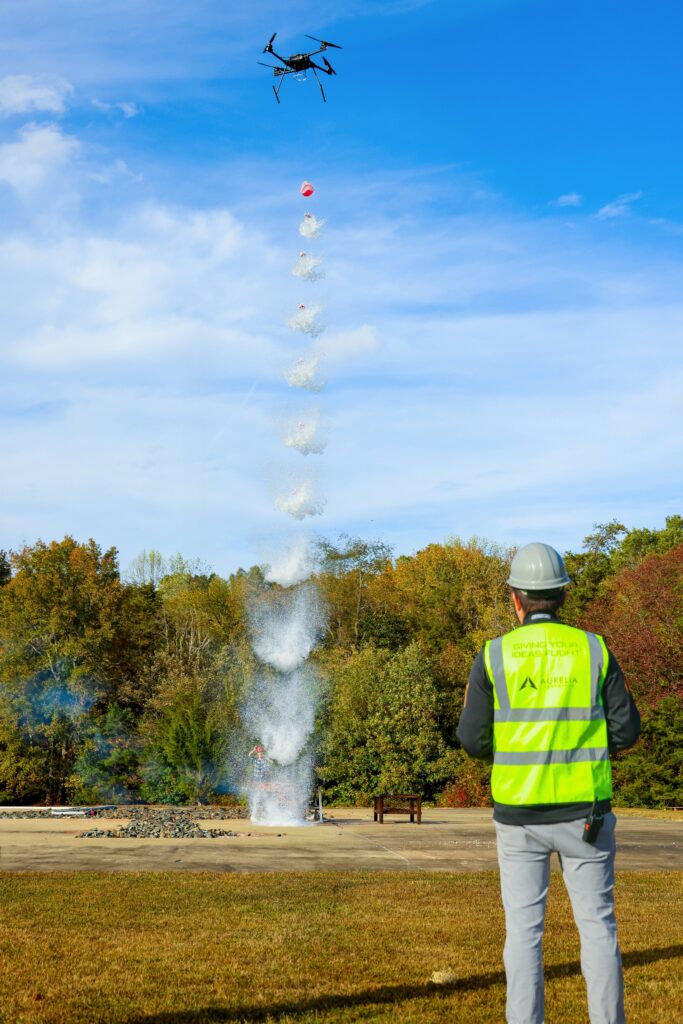 A Man In A Hard Hat And High Visibility Vest Looks On As A Drone Drops Water To Extinguish A Fire Scaled